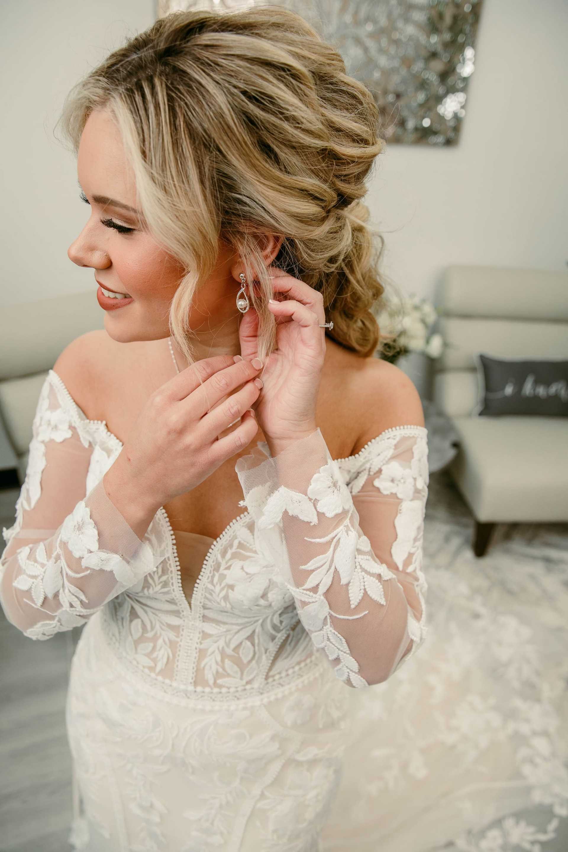 Bride in lace wedding gown adjusts her earring, smiling in a softly lit room.